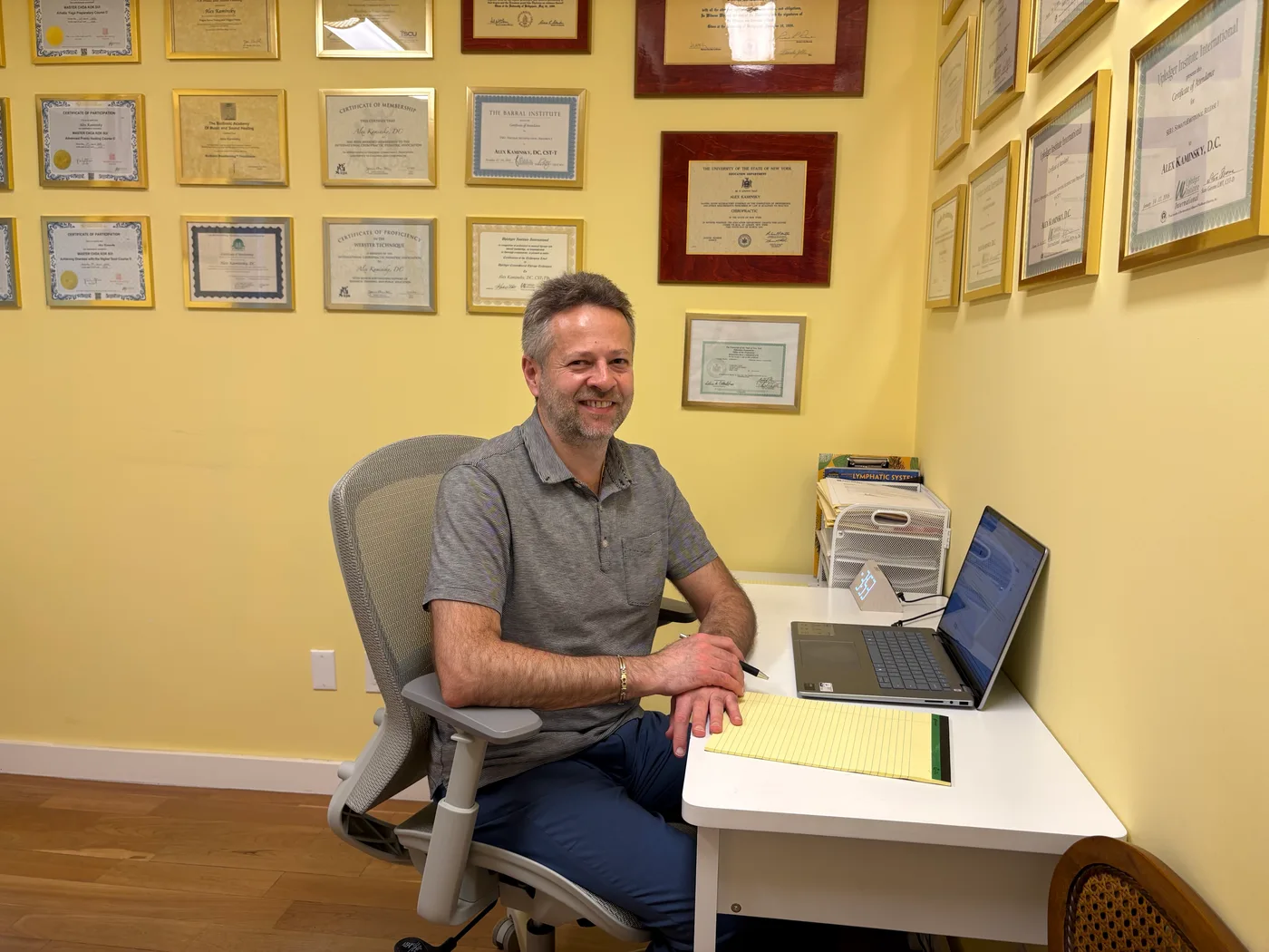 Dr. Alex Kaminsky at his desk in his Midtown Manhattan office, with his framed craniosacral therapy certifications on the wall behind him.