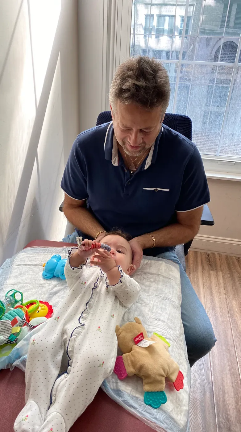 Infant lying on the treatment table with toys while Dr. Kaminsky performs craniosacral therapy by a sunlit Manhattan window.